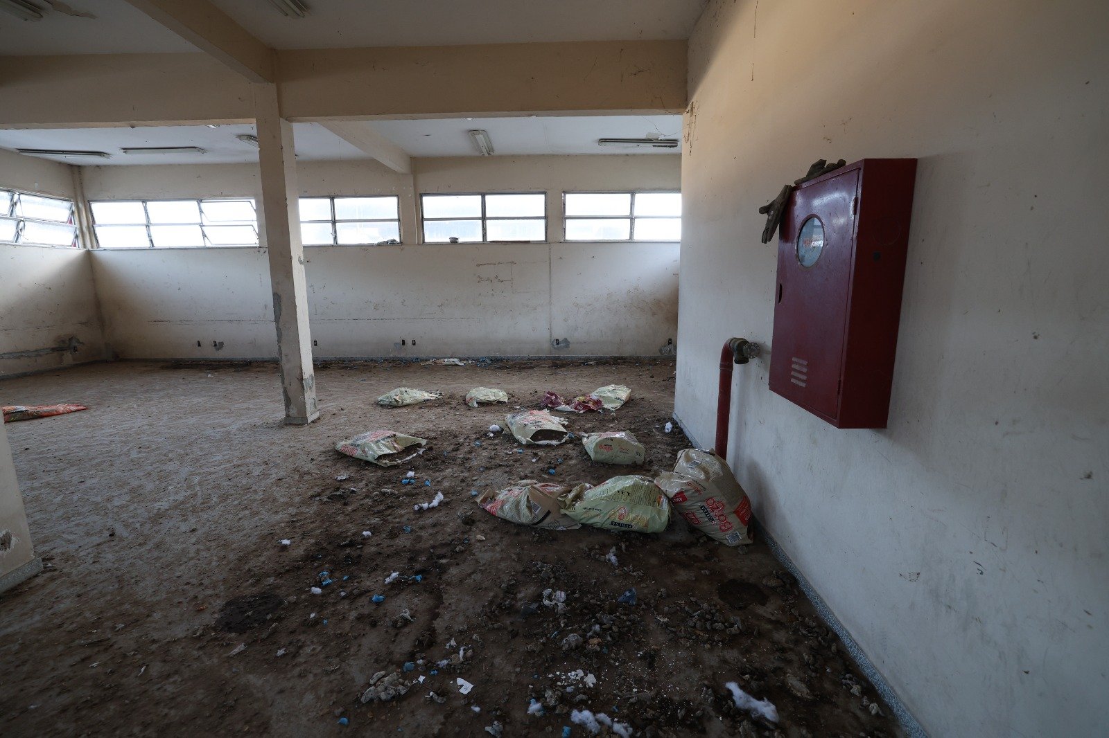 Sala vazia com piso coberto de terra, entulho e sacos de ração, ao lado de um hidrante, no imóvel ocupado pela Brigada K9