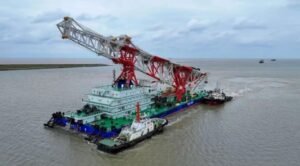 Navio de cravação de estacas Tie Jian Da Qiao Zhuang 1, com guindaste vermelho e branco, rebocado por embarcações de apoio ao deixar o Porto de Lüsi, na China