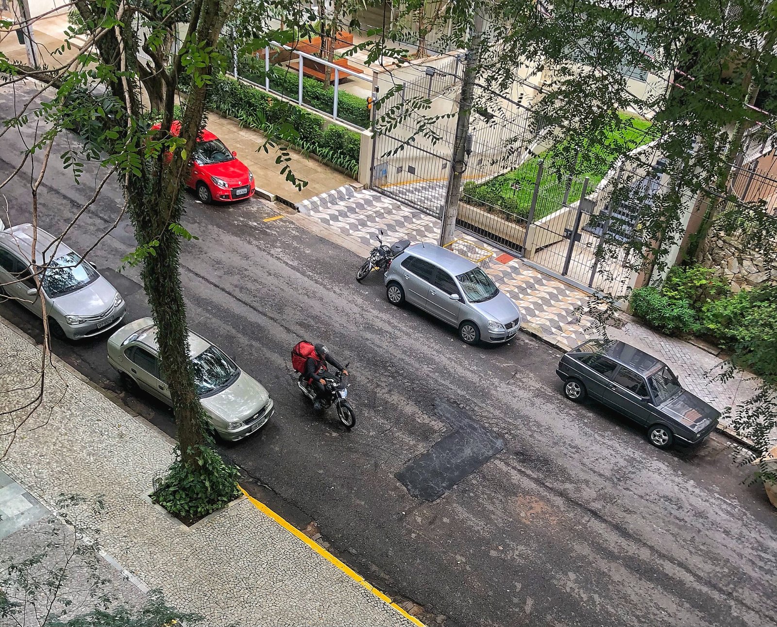Motociclista de capacete trafega entre carros estacionados em rua, vista de cima